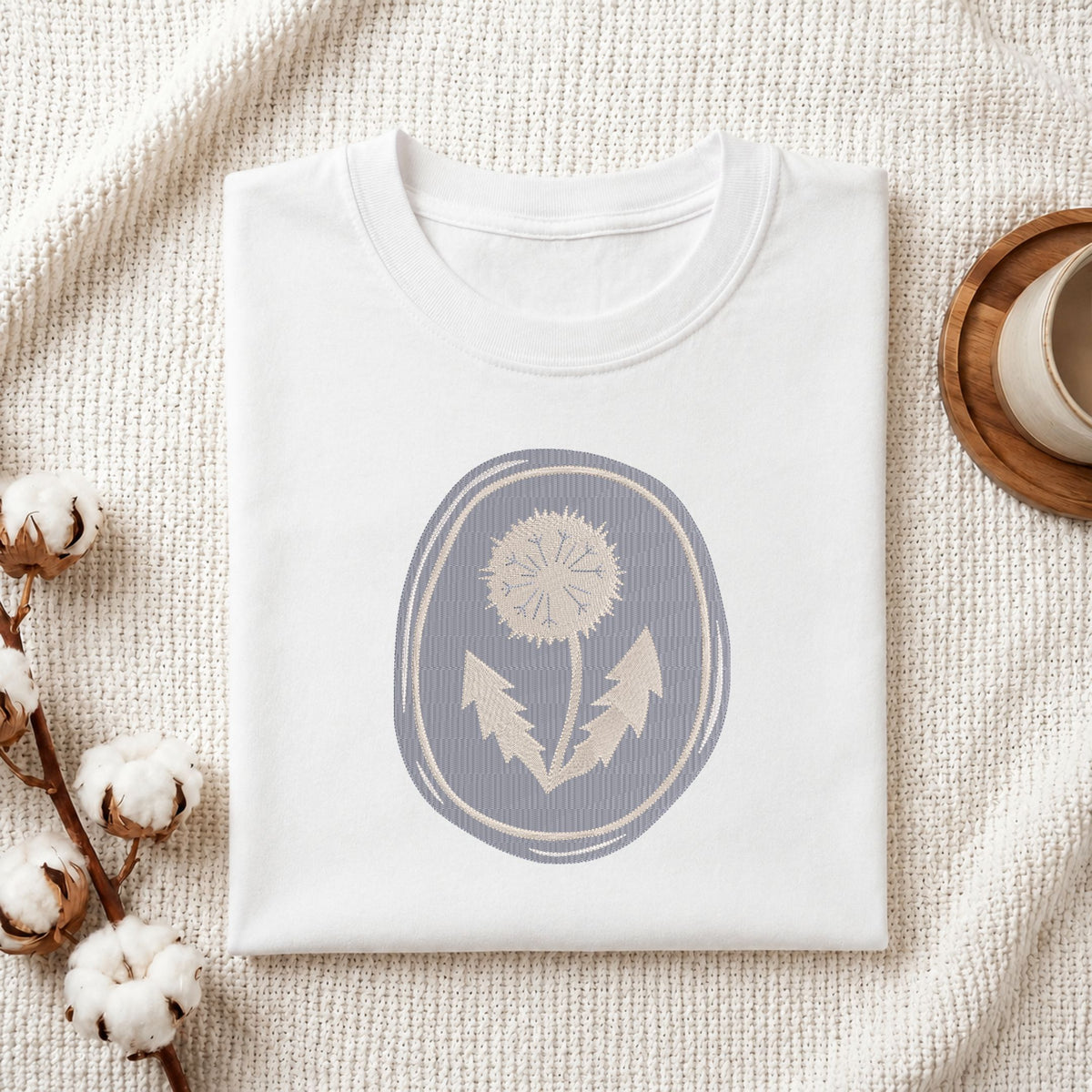 A folded white crewneck t-shirt with a muted blue-gray oval graphic of a dandelion, styled on a textured cream knit blanket with cotton bolls and a wooden cup in the background.