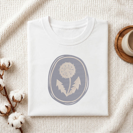 A folded white crewneck t-shirt with a muted blue-gray oval graphic of a dandelion, styled on a textured cream knit blanket with cotton bolls and a wooden cup in the background.