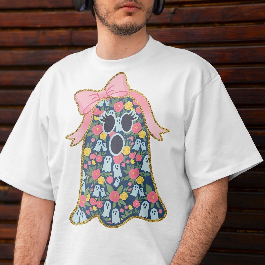 A man wearing a white crewneck t-shirt with a colorful graphic of a cute ghost character decorated with flowers and a pink bow, set against a dark wooden background.