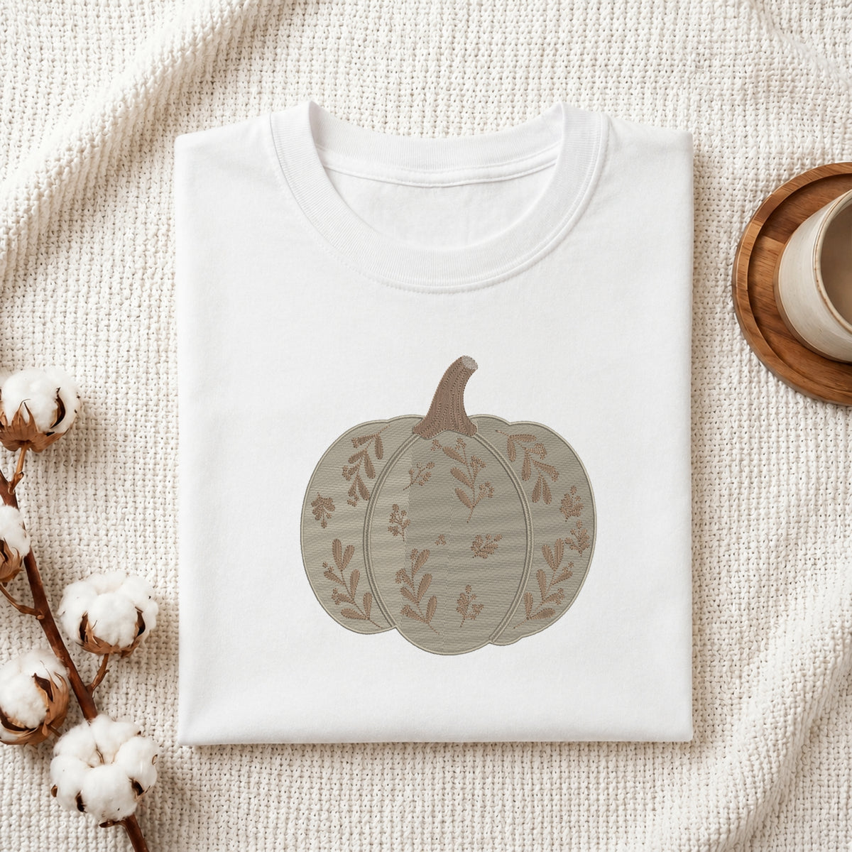 A folded white crewneck t-shirt featuring a printed autumn pumpkin design in muted beige and brown tones, styled on a textured cream fabric with cotton bolls and a wooden cup in the background.