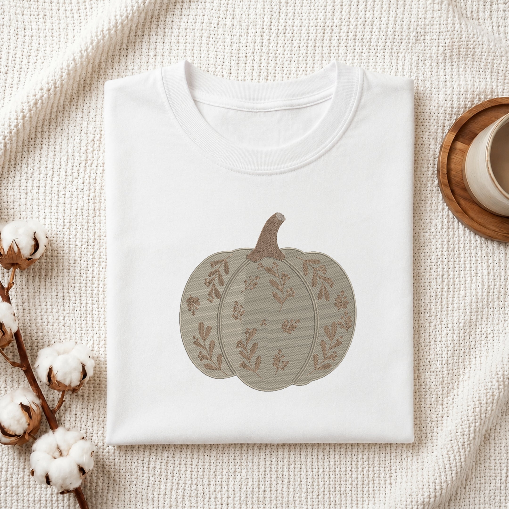 A folded white crewneck t-shirt featuring a printed autumn pumpkin design in muted beige and brown tones, styled on a textured cream fabric with cotton bolls and a wooden cup in the background.