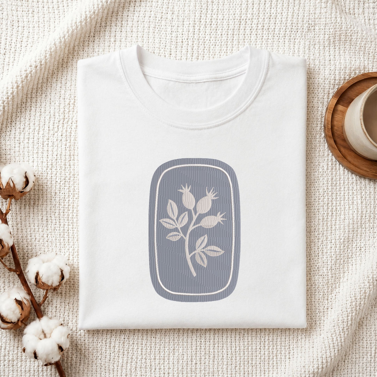 Folded white cotton t-shirt mockup featuring a minimalist botanical rosehip machine embroidery design, styled with cotton stems and a wooden cup.