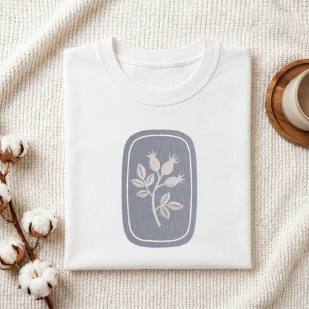 Folded white cotton t-shirt mockup featuring a minimalist botanical rosehip machine embroidery design, styled with cotton stems and a wooden cup.