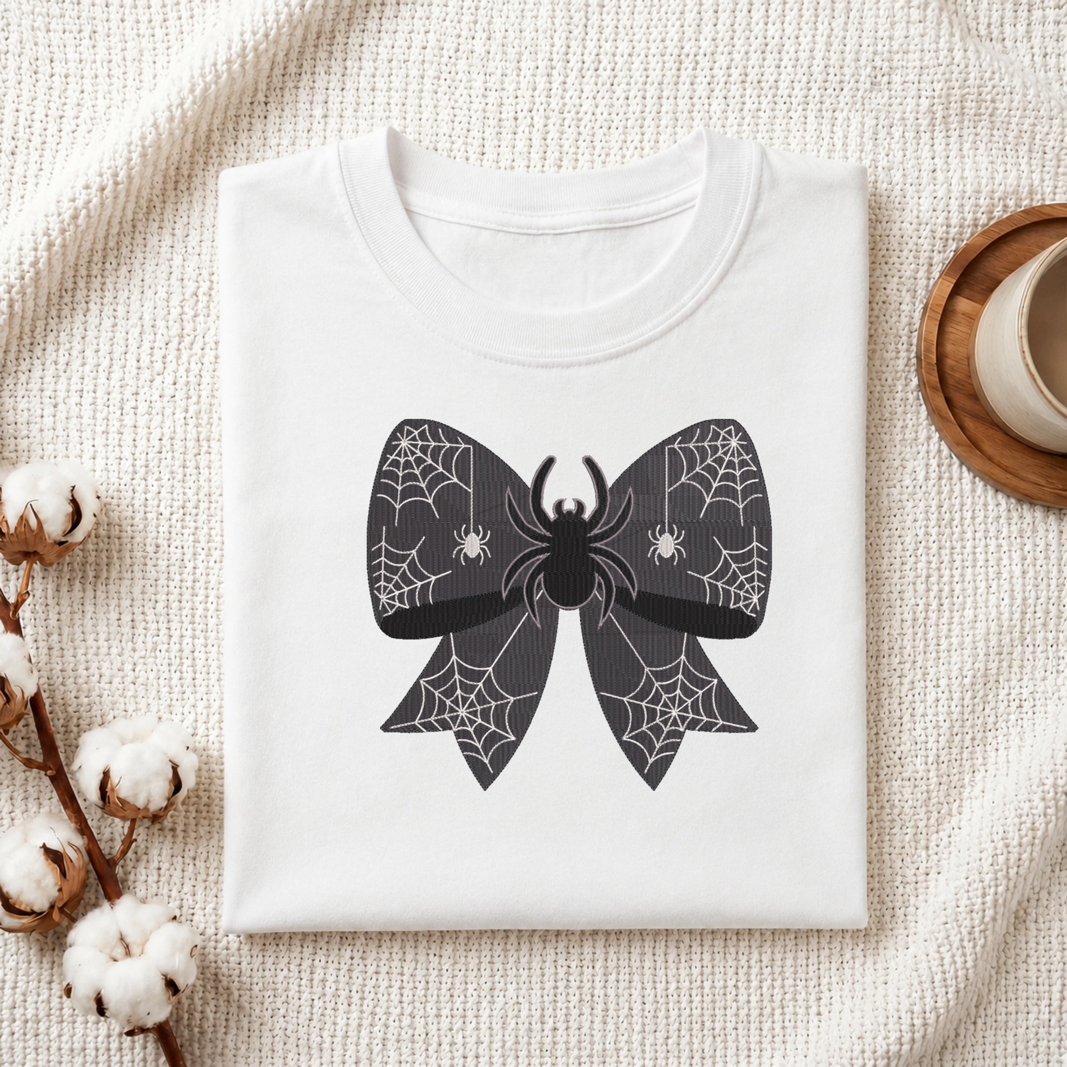 A neatly folded white t-shirt featuring the embroidered spider web bow design, displayed on a textured white knit background with cotton stems and a wooden cup.
