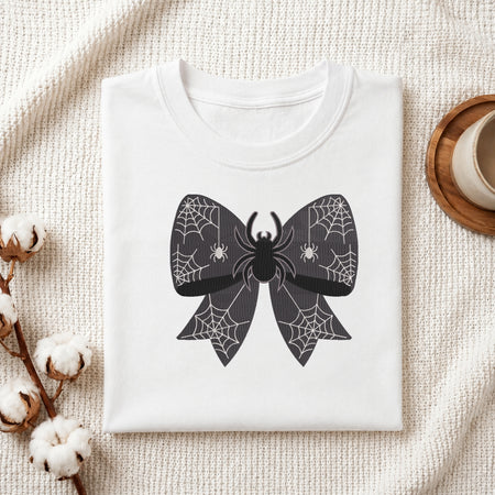 A neatly folded white t-shirt featuring the embroidered spider web bow design, displayed on a textured white knit background with cotton stems and a wooden cup.