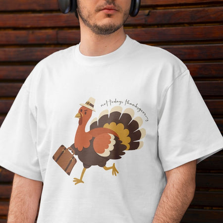 A man wearing a white crewneck t-shirt with a graphic of a cartoon turkey carrying a suitcase and wearing a hat, with the text 'not today, thanksgiving' written above it. The man is wearing black headphones against a dark brown wooden background.