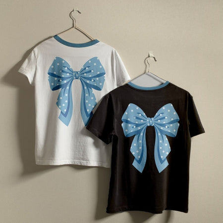 Two short-sleeve ringer T-shirts hanging on hangers against a plain beige wall, showing the back designs: one white shirt with a light blue bow with white polka dots, and one black shirt with the same light blue bow design.