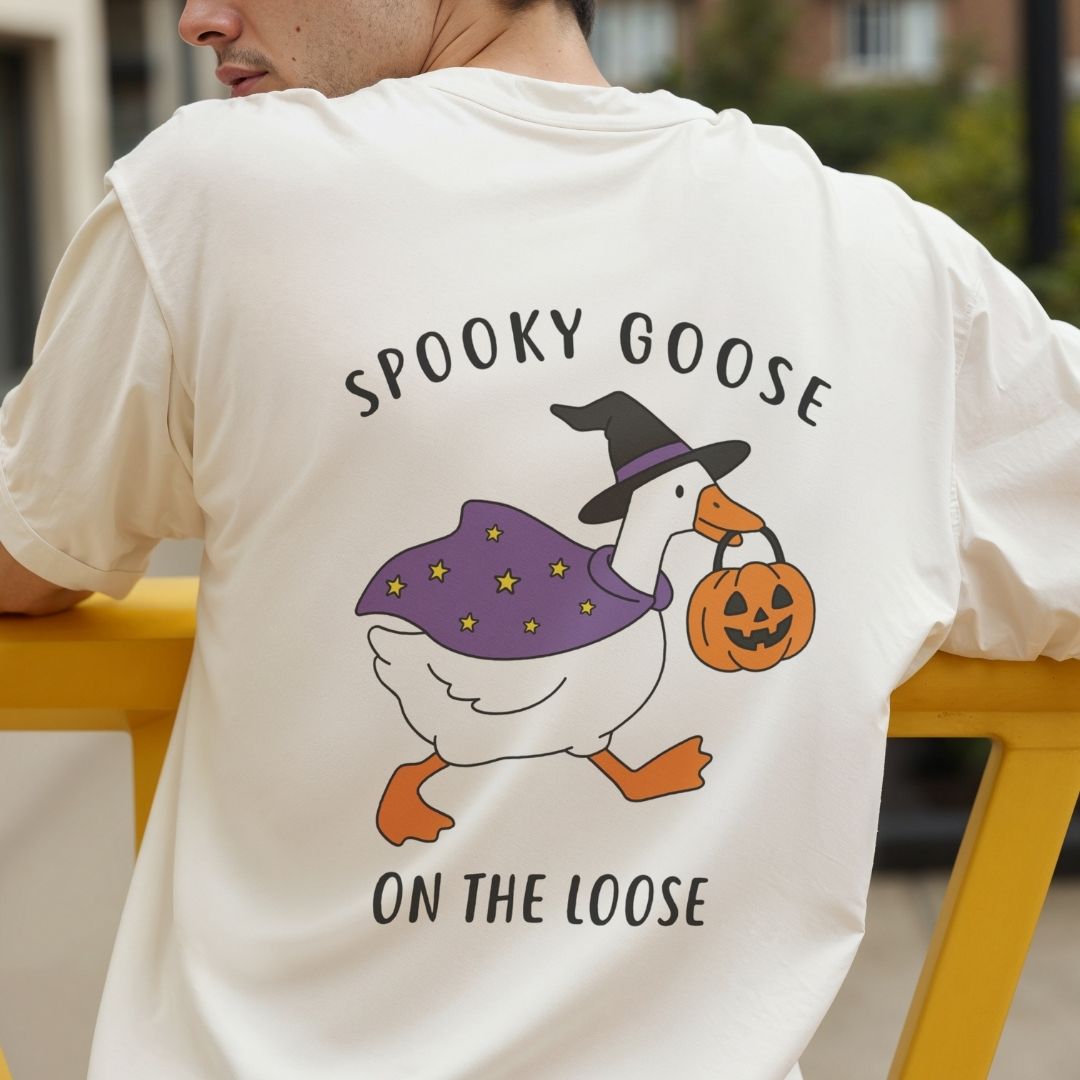 A person wearing a cream-colored t-shirt viewed from the back, showcasing a large Spooky Goose on the Loose witch design centered on the back.