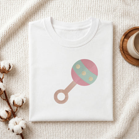 A folded white baby t-shirt with a pastel pink and mint green rattle graphic, styled on a textured cream knit blanket with cotton bolls and a wooden cup.