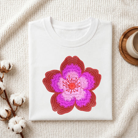 A folded white crewneck t-shirt featuring a large, stylized embroidered cherry blossom design in shades of red, pink, and purple. The t-shirt is styled in a flat-lay arrangement on a textured cream knit blanket, accompanied by cotton bolls on the left and a wooden cup on a coaster on the right.