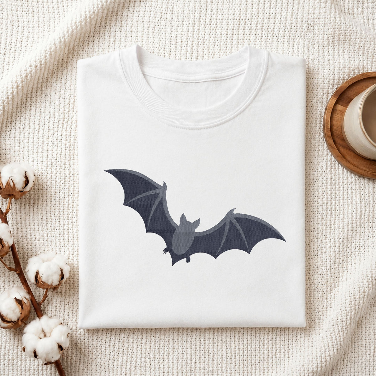 A folded white crewneck t-shirt featuring a dark gray cartoon bat graphic on the front, styled on a textured cream knit blanket with cotton bolls and a wooden cup in the background.