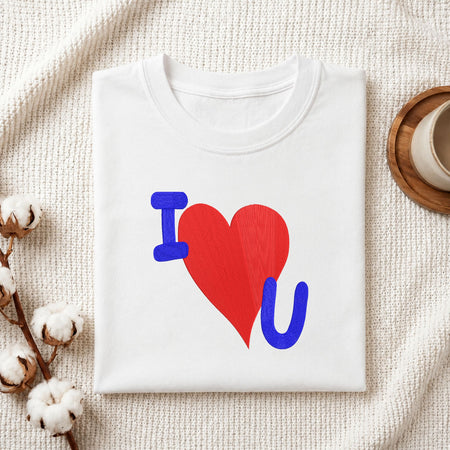 Folded white cotton t-shirt mockup featuring an "I Love You" machine embroidery design, styled with cotton stems and a wooden cup