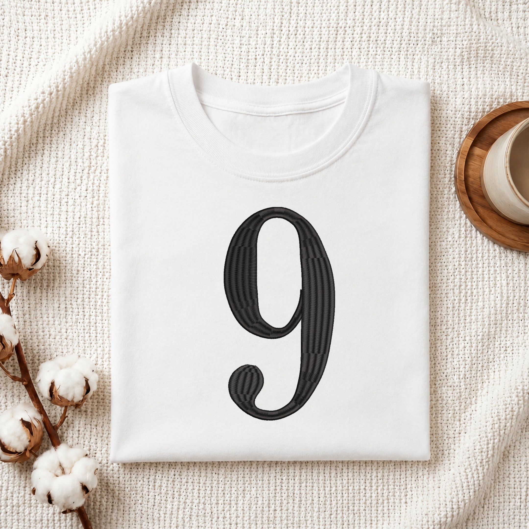 A folded white T-shirt with a large black decorative numeral '9' printed on the front, styled on a textured cream fabric with cotton stems and a small cup in the corners.