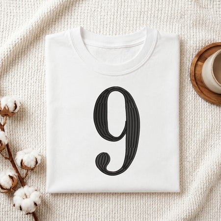 A folded white T-shirt with a large black decorative numeral '9' printed on the front, styled on a textured cream fabric with cotton stems and a small cup in the corners.