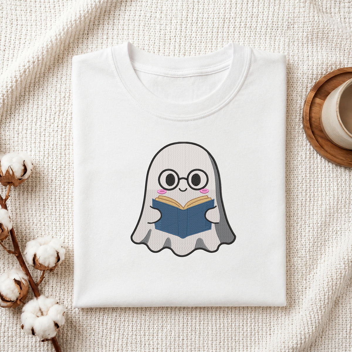 A folded white crewneck t-shirt featuring a cute cartoon ghost character with glasses reading a blue book, styled on a textured cream knit blanket with cotton bolls and a wooden cup in the background.