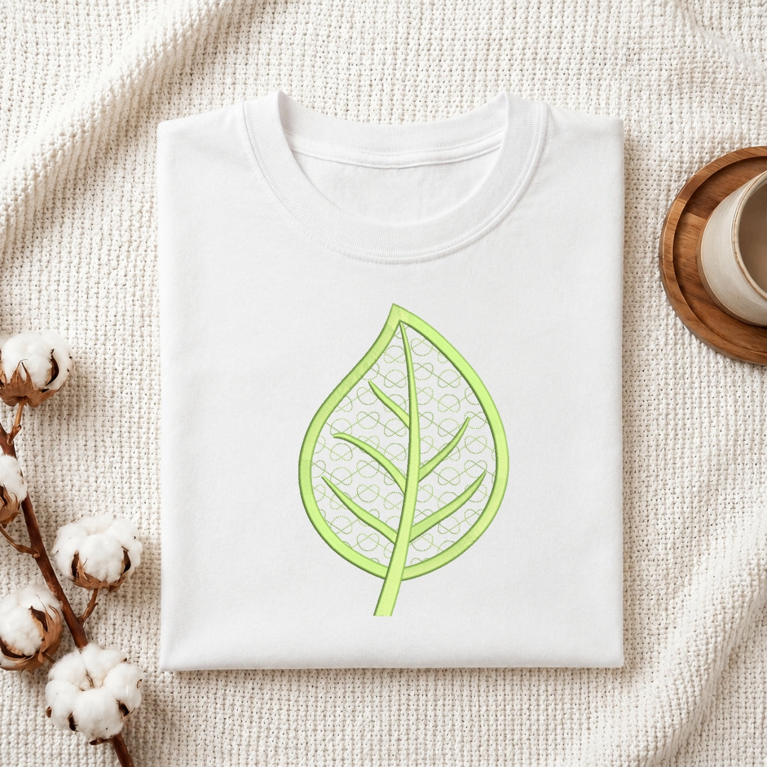 A folded white crewneck t-shirt with a light green leaf graphic printed on the chest, styled on a textured cream knit blanket with cotton bolls and a wooden cup in the background.