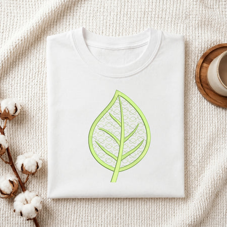 A folded white crewneck t-shirt with a light green leaf graphic printed on the chest, styled on a textured cream knit blanket with cotton bolls and a wooden cup in the background.