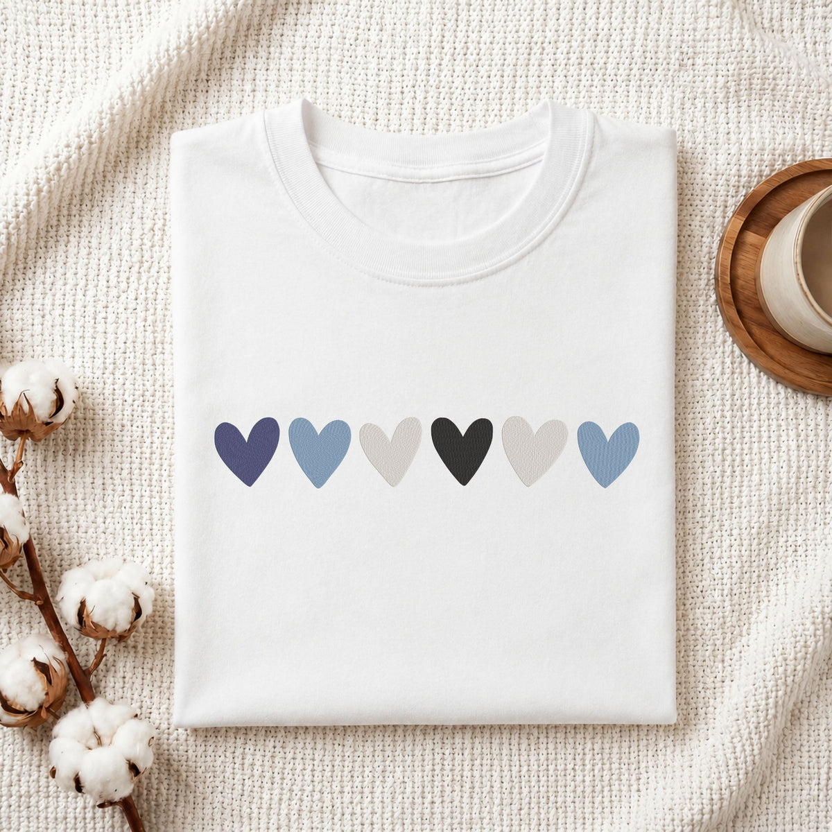 A folded white crewneck t-shirt with a row of six textured heart shapes in shades of blue, gray, and black across the chest, displayed on a textured cream knit blanket with cotton bolls and a wooden cup as props.