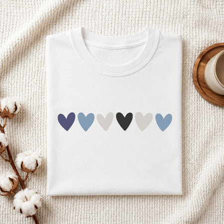 A folded white crewneck t-shirt with a row of six textured heart shapes in shades of blue, gray, and black across the chest, displayed on a textured cream knit blanket with cotton bolls and a wooden cup as props.
