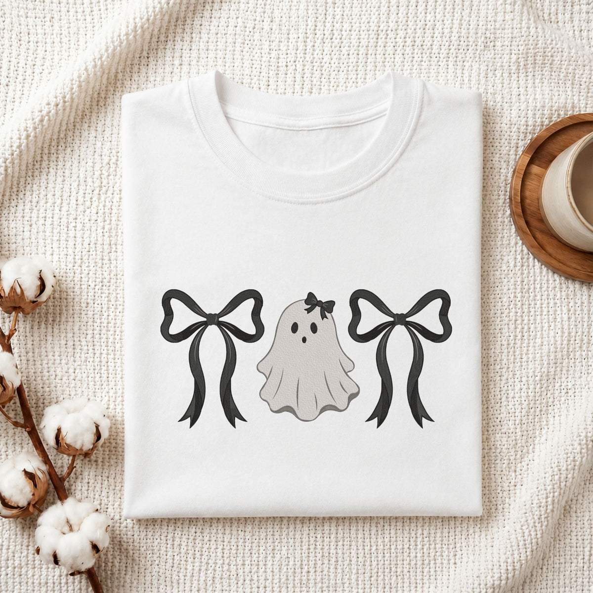  Folded white cotton t-shirt mockup featuring a cute ghost with bows machine embroidery design, styled with cotton stems and a wooden cup   