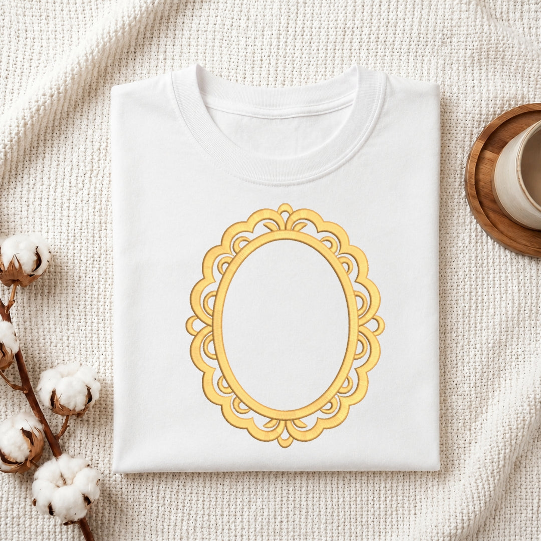 A folded white t-shirt with a gold ornate oval frame graphic on the front, styled on a textured cream fabric background with cotton bolls and a wooden cup.