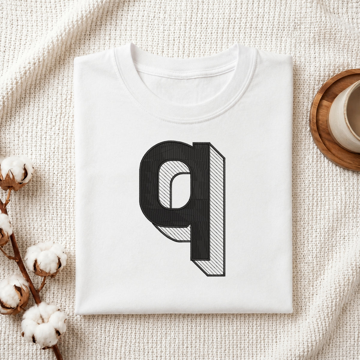 A folded white t-shirt with a large black 3D-effect letter 'q' graphic on the front, styled on a textured cream fabric with cotton bolls and a wooden cup.