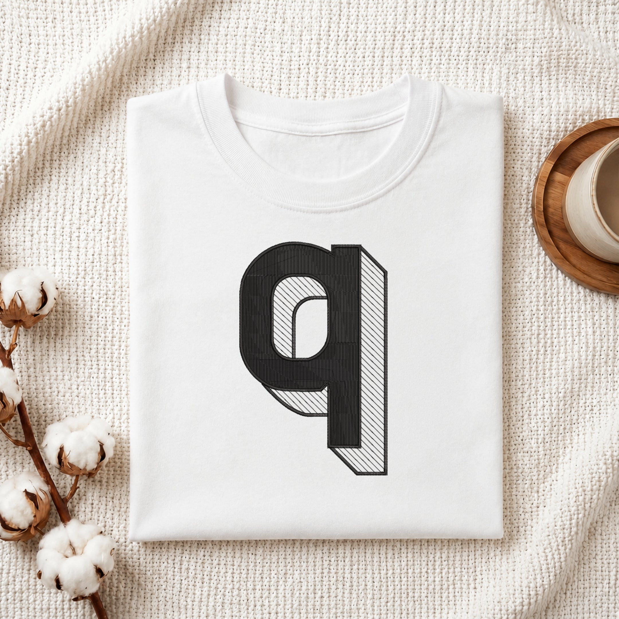 A folded white t-shirt with a large black 3D-effect letter 'q' graphic on the front, styled on a textured cream fabric with cotton bolls and a wooden cup.