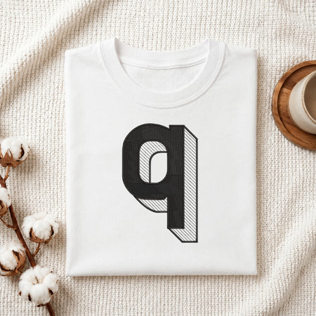 A folded white t-shirt with a large black 3D-effect letter 'q' graphic on the front, styled on a textured cream fabric with cotton bolls and a wooden cup.