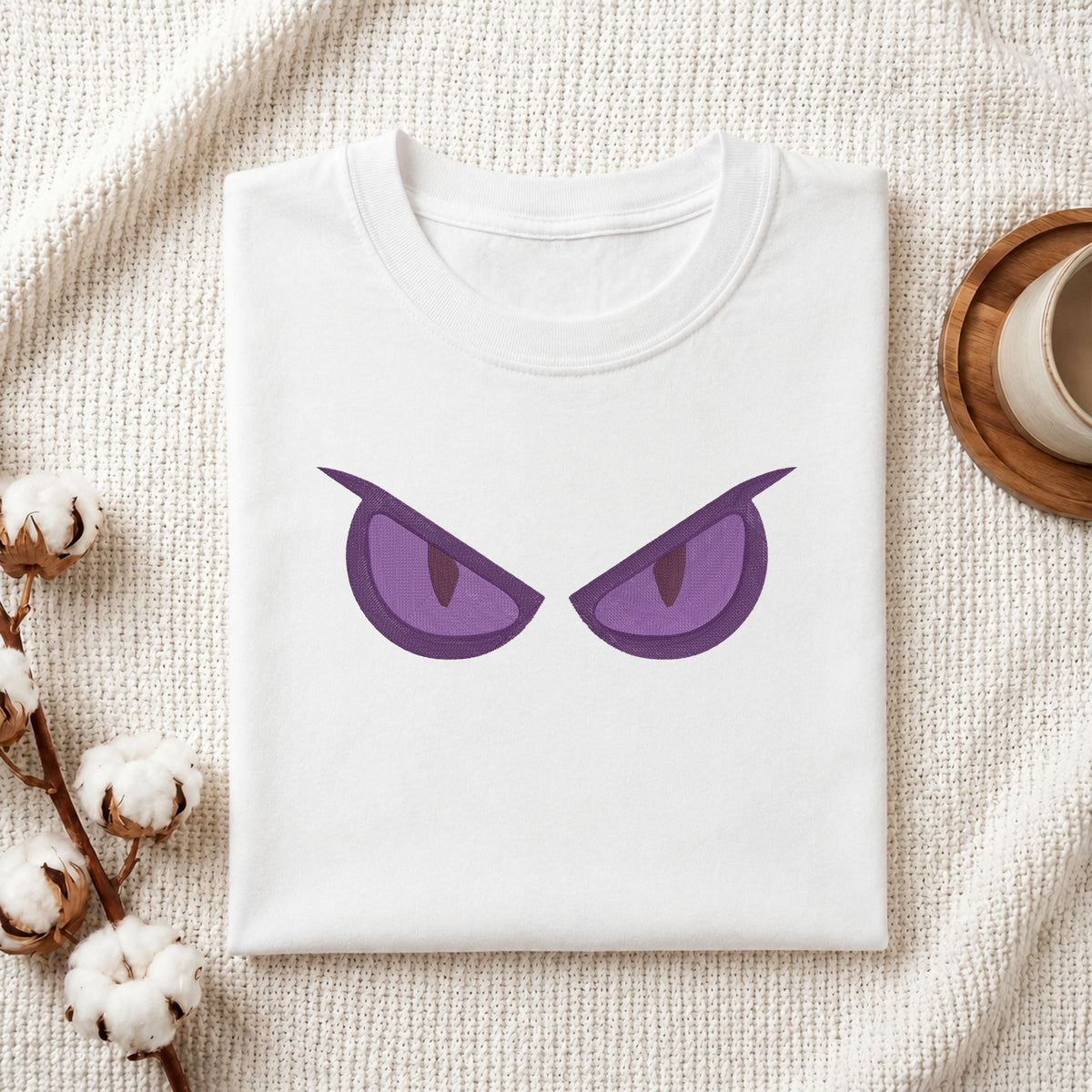 A folded white t-shirt with a graphic of two purple angry eyes, styled on a textured cream knit blanket with cotton bolls and a cup on a wooden tray.