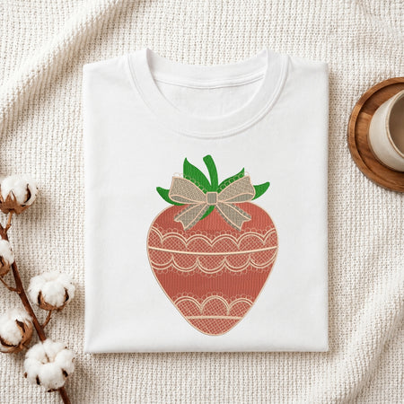 A folded white crewneck t-shirt with a decorative embroidered strawberry design, styled on a textured cream fabric with cotton bolls and a wooden cup.