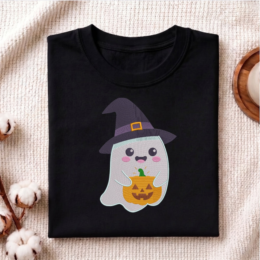 A folded black t-shirt with a cute cartoon ghost wearing a purple witch hat and holding a jack-o'-lantern pumpkin, displayed on a textured beige fabric background with cotton bolls and a wooden coaster.