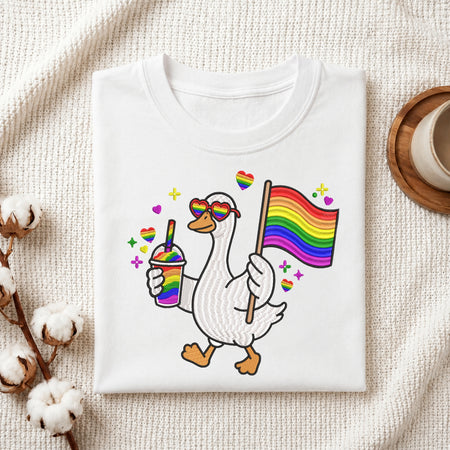 A folded white t-shirt with a colorful graphic of a walking goose wearing rainbow heart sunglasses, holding a rainbow flag and a rainbow drink, styled on a textured cream fabric with cotton bolls and a wooden cup.