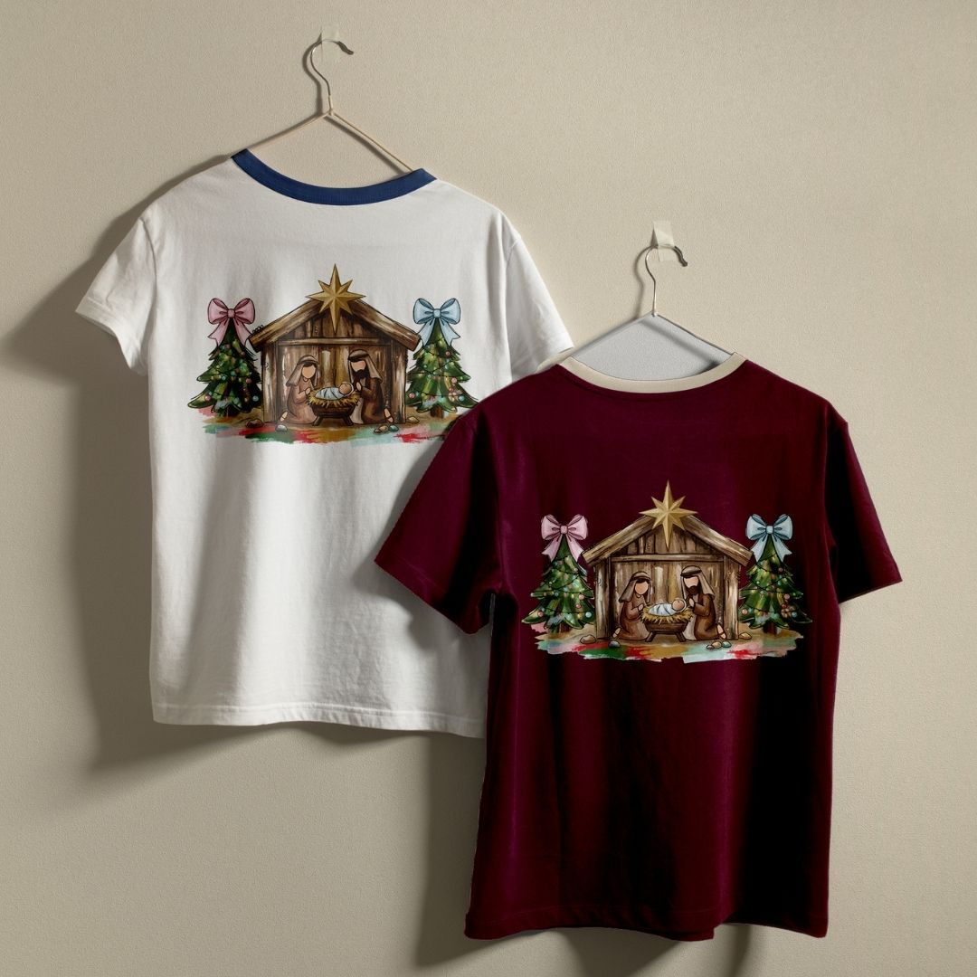 Two Christmas-themed ringer t-shirts hanging on hangers against a plain beige wall. The left shirt is white with a blue collar and features a Nativity scene printed on the back.
