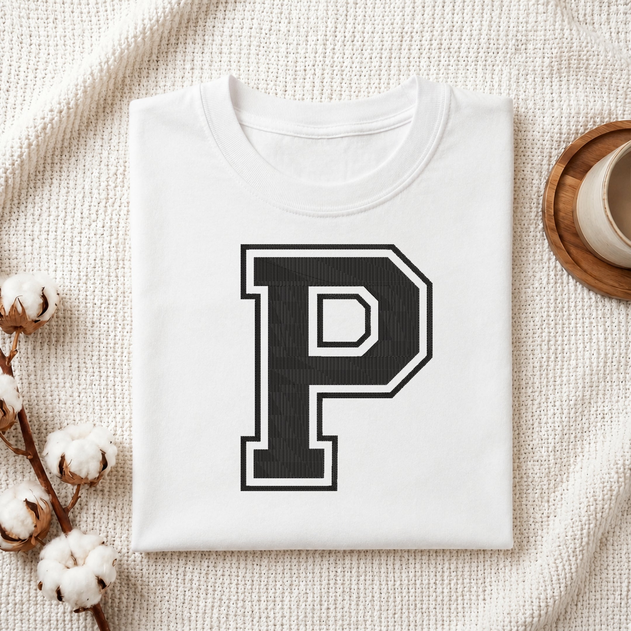 A folded white crewneck t-shirt with a large black and white block letter 'P' printed on the chest, styled on a textured cream knit blanket with cotton bolls and a wooden cup in the background.