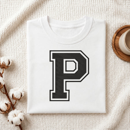 A folded white crewneck t-shirt with a large black and white block letter 'P' printed on the chest, styled on a textured cream knit blanket with cotton bolls and a wooden cup in the background.