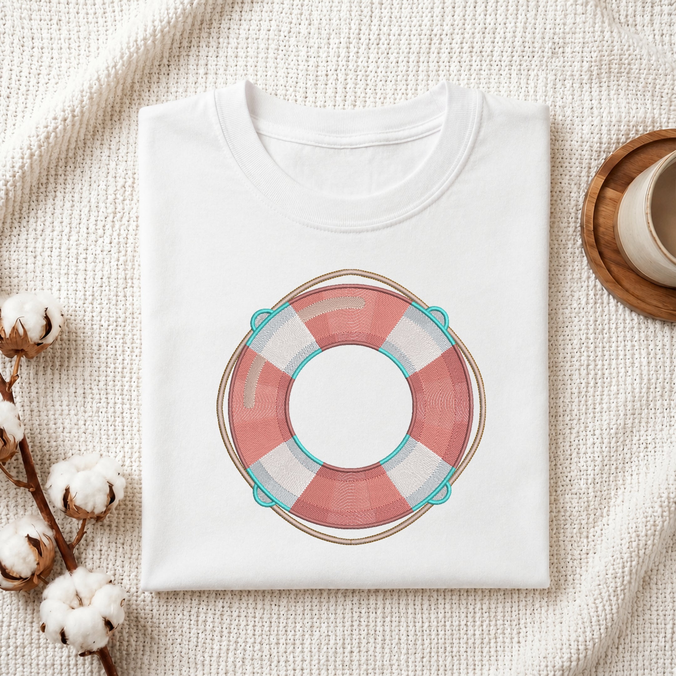 A folded white crewneck t-shirt with a pink and white striped life preserver graphic, styled on a textured cream fabric with cotton bolls and a wooden cup.