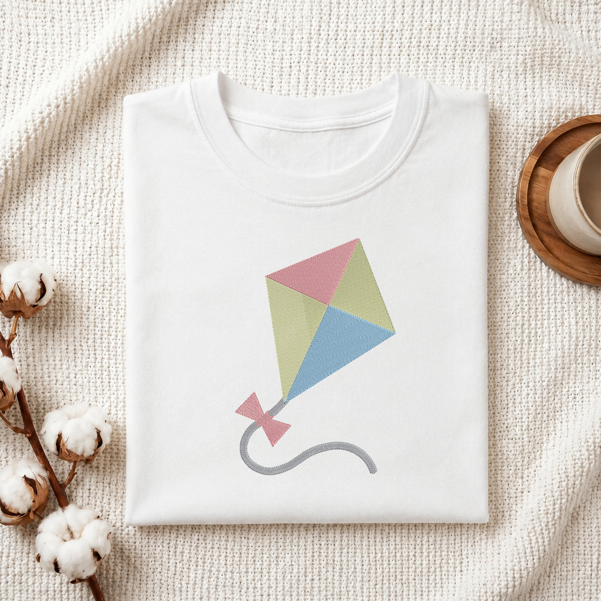 A neatly folded white crewneck t-shirt featuring a stitched colorful kite design, styled with cotton branches and a wooden coaster.