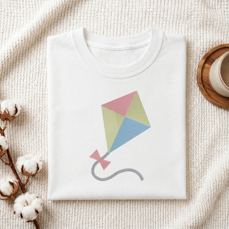 A neatly folded white crewneck t-shirt featuring a stitched colorful kite design, styled with cotton branches and a wooden coaster.