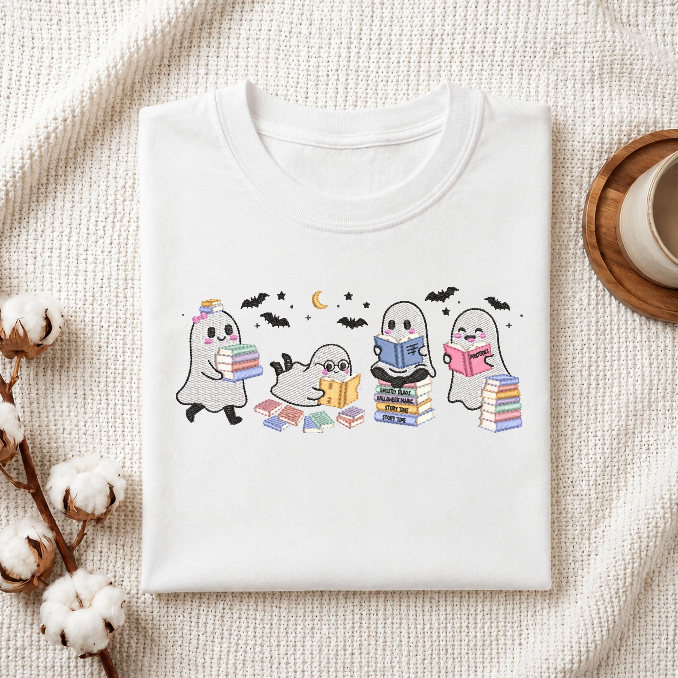 A folded white graphic T-shirt displayed on a textured cream fabric background, styled with cotton bolls and a cup on a wooden coaster. The shirt features a cute illustration of three ghosts reading books under a crescent moon.