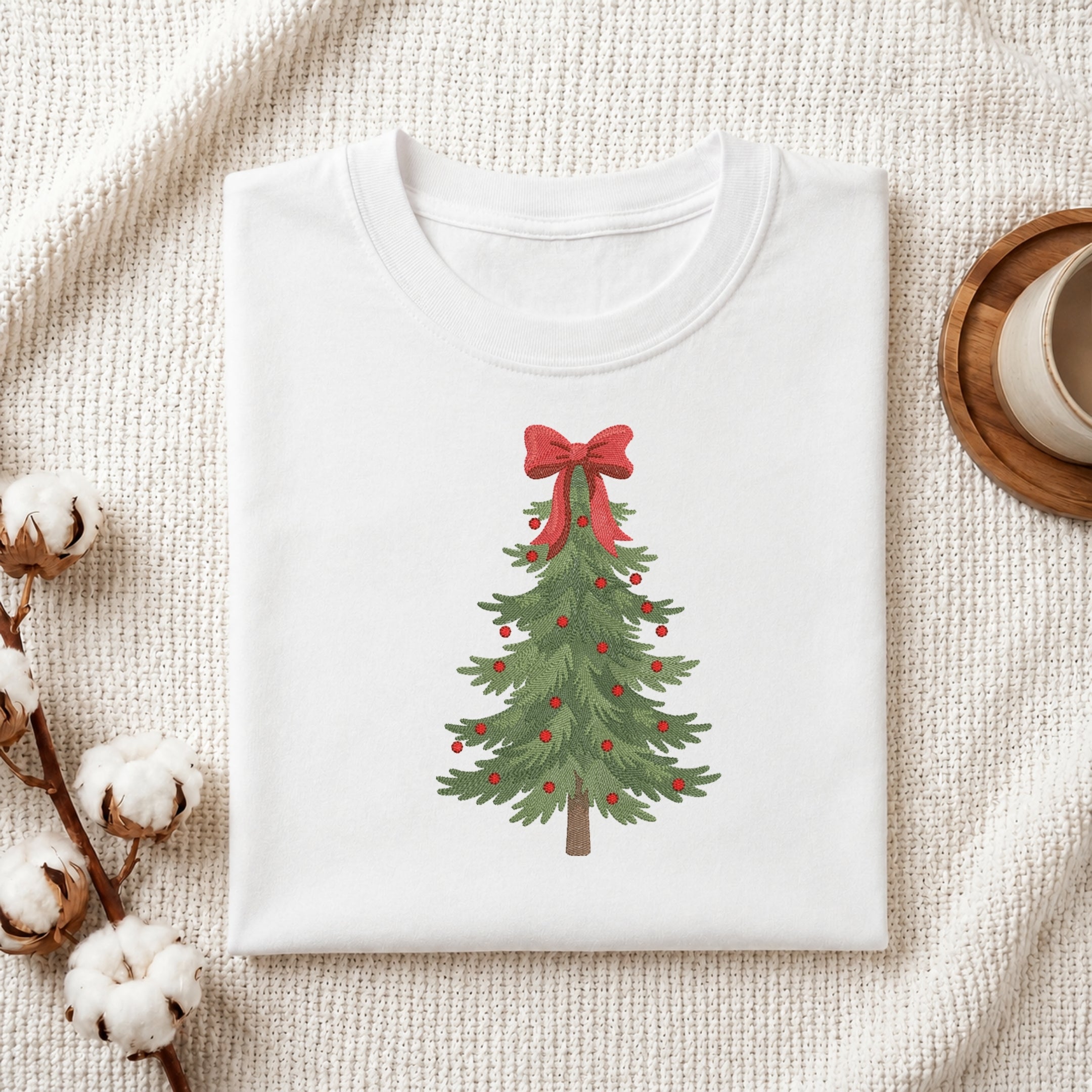 A folded white crewneck t-shirt with a watercolor Christmas tree graphic, styled on a textured cream fabric with cotton bolls and a wooden mug.