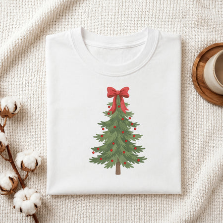 A folded white crewneck t-shirt with a watercolor Christmas tree graphic, styled on a textured cream fabric with cotton bolls and a wooden mug.