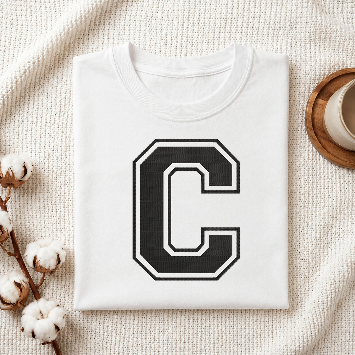 A folded white T-shirt with a large black block letter 'C' outlined in white on the front, styled on a textured cream fabric with cotton stems and a small cup at the side.