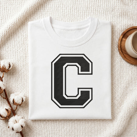 A folded white T-shirt with a large black block letter 'C' outlined in white on the front, styled on a textured cream fabric with cotton stems and a small cup at the side.