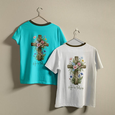 Two t-shirts hanging on hangers, one turquoise and one white, both printed with a floral cross graphic and the text "He is Resign".