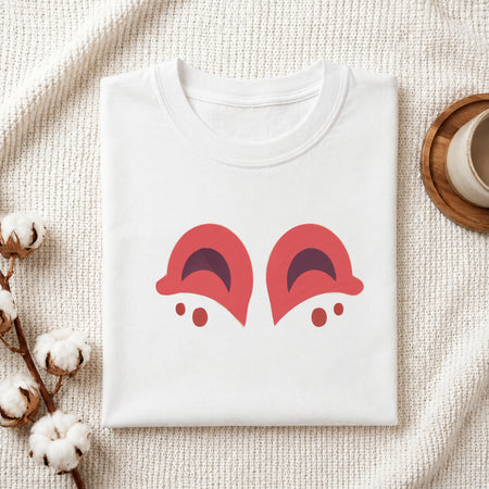 A folded white t-shirt with a graphic of two red cartoon eyes on the front, styled in a flat lay on a textured cream fabric with cotton bolls and a wooden cup.