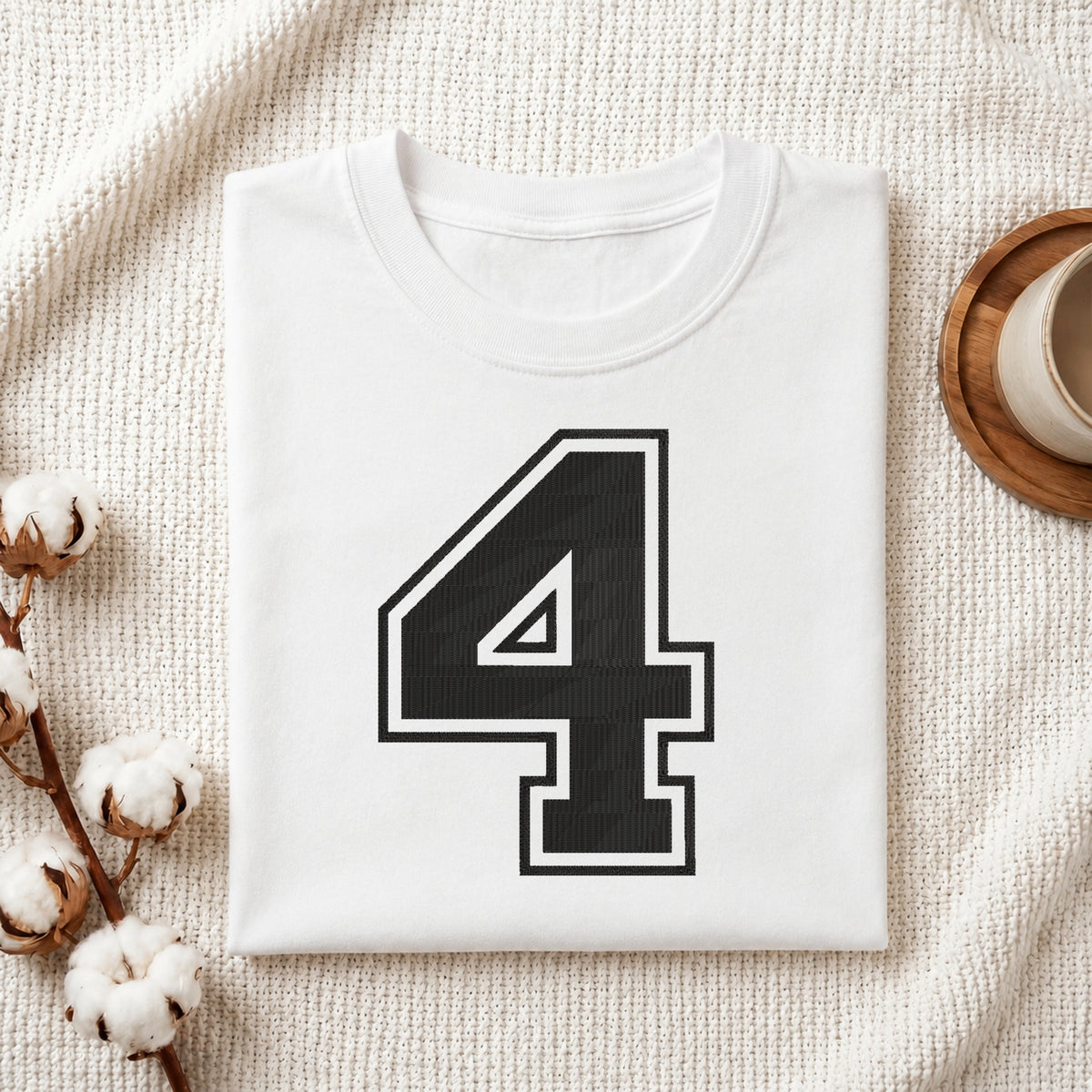 A folded white t-shirt with a large black number 4 outlined in white on the front, styled on a textured cream fabric with cotton bolls and a wooden cup.