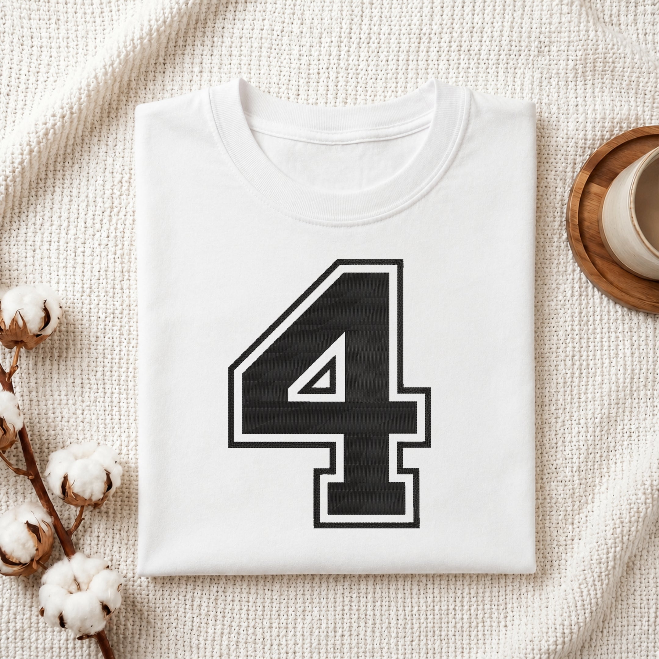 A folded white t-shirt with a large black number 4 outlined in white on the front, styled on a textured cream fabric with cotton bolls and a wooden cup.