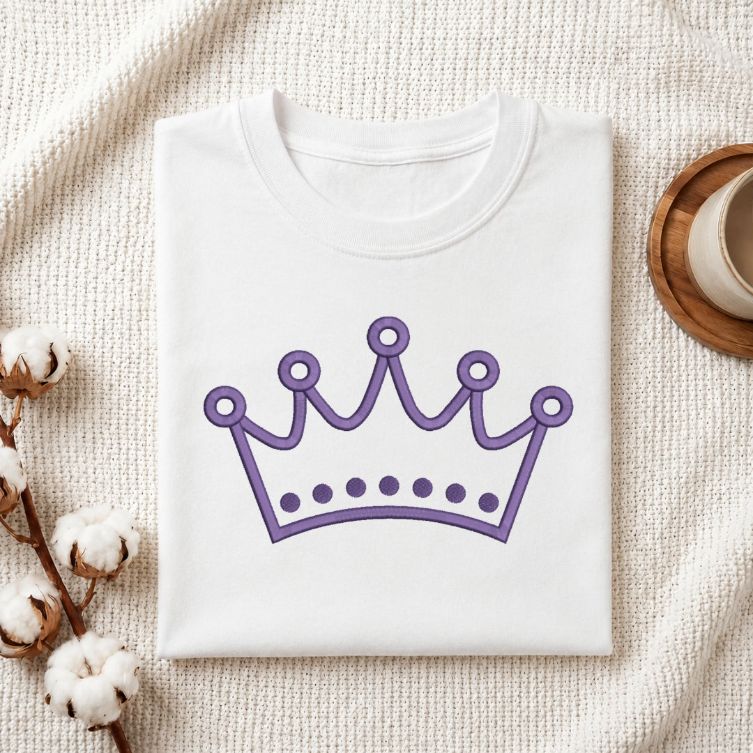 A folded white crewneck t-shirt with a purple crown graphic printed on the front, styled on a textured cream knit blanket with cotton bolls and a wooden cup in the background.