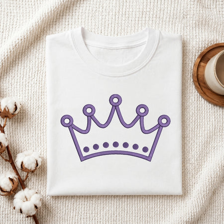 A folded white crewneck t-shirt with a purple crown graphic printed on the front, styled on a textured cream knit blanket with cotton bolls and a wooden cup in the background.