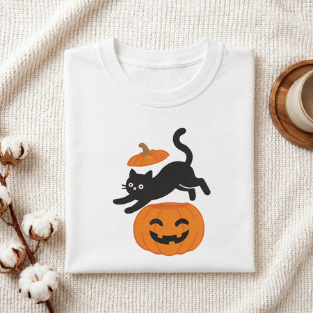 Folded white cotton t-shirt mockup featuring a playful black cat jumping over a jack-o-lantern pumpkin machine embroidery design, styled with cotton stems and a wooden cup   